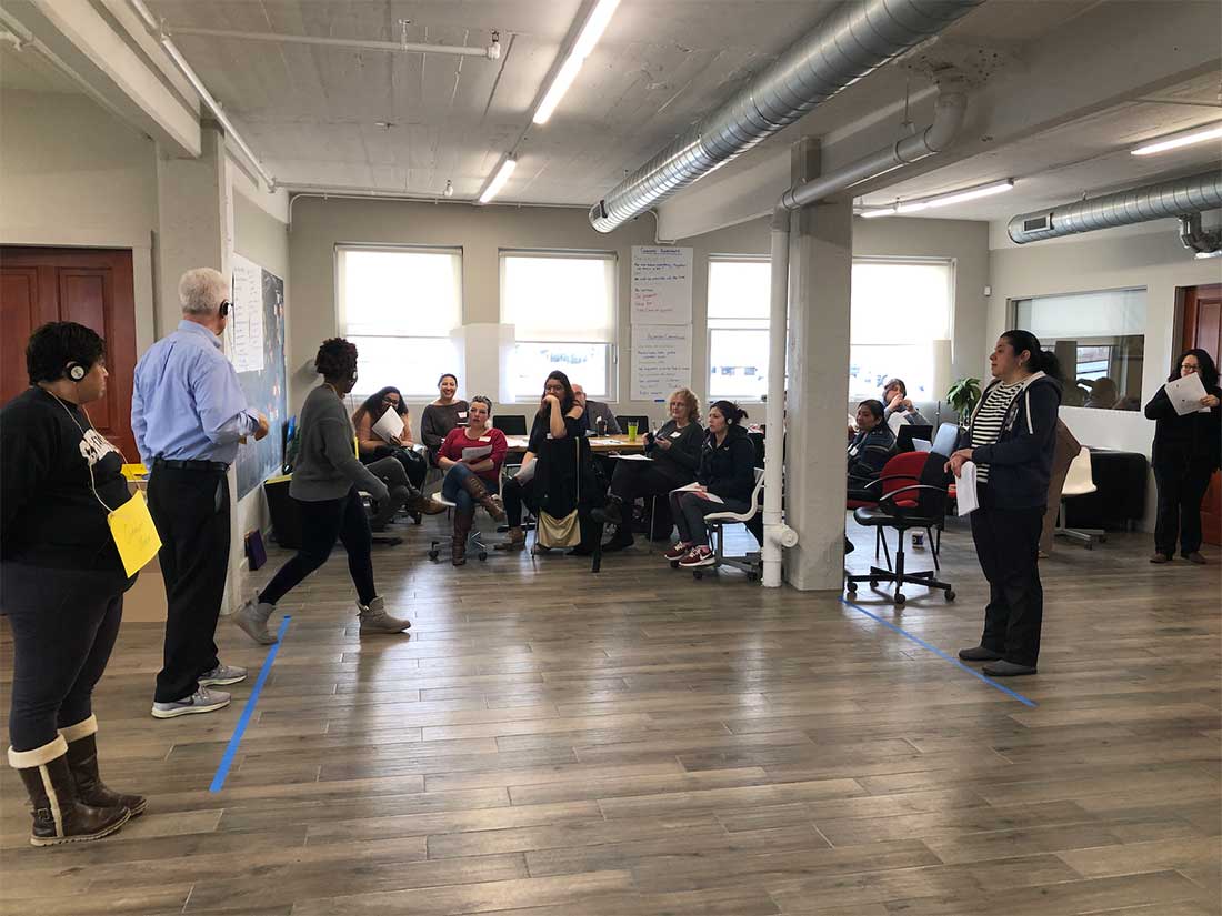 Una fotografía que muestra personas en un grupo grande durante el taller “¡Convénceme!” La sala tiene dos líneas de cinta azul en el piso, y un grupo de los/las/les participantes está detrás de una línea, frente a otro/a/e participantes que está detrás de la segunda línea y está hablando con él/ella/elle.
